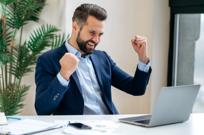 Successful joyful lucky caucasian businessman in a suit, ceo company, manager, sits at work desk in the office, gesturing with fists, celebrate success, win, big profit, good deal. Victory concept